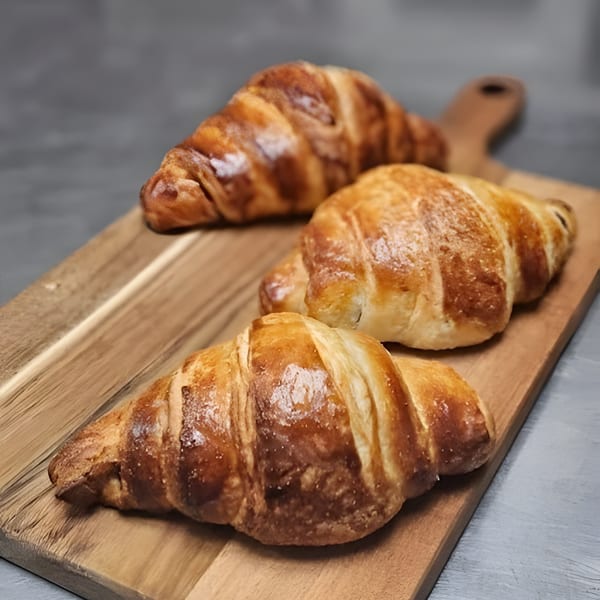 Three golden-brown croissants with flaky crusts rest on a rustic wooden cutting board.